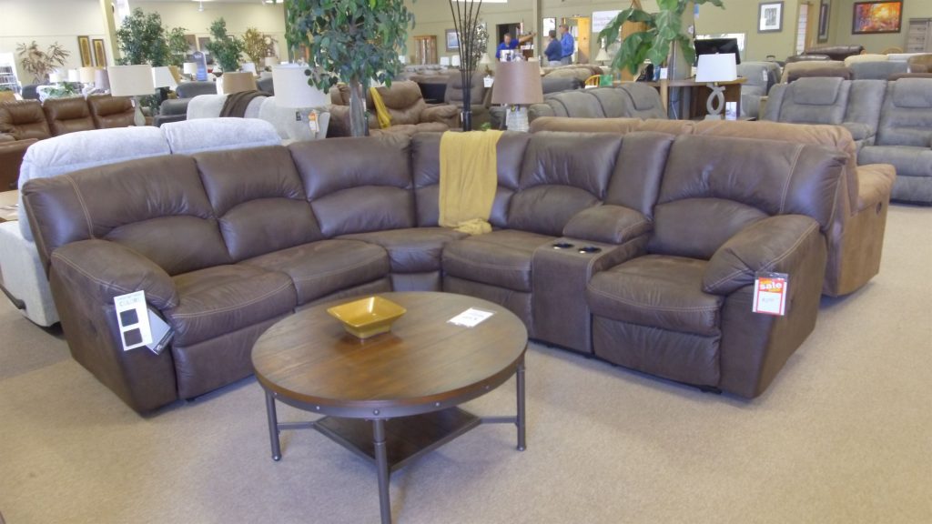 Living room furniture display; Dark brown sectional couch in showroom, with built in cup holders and reclinig ends, draped with a yellow blanket in the center. ROund coffee table with small bowl in front of couch.